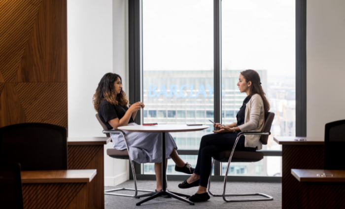 Two women sat opposite each other over a table discussing work in a skyscraper building with a view of London in the background